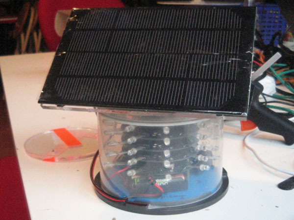 Round plastic enclosure with a number of LEDs visible inside, on top of a battery pack. A solar panel site on top of the round enclosure.